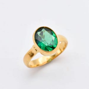 May include: A gold ring featuring a large, oval-cut, faceted green gemstone set in a smooth, polished gold band. The vibrant green stone is securely held within a simple bezel setting, highlighting the clear, reflective quality of the gem.