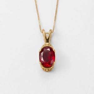May include: A gold pendant necklace featuring an oval-cut deep red ruby gemstone set in a yellow gold frame with intricate metalwork details at the base, suspended from a delicate matching gold chain against a plain white background.