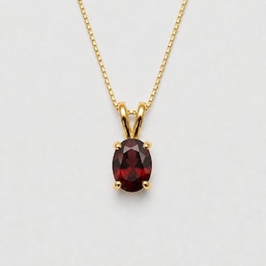 May include: A gold necklace featuring an oval-cut deep red garnet gemstone pendant. The stone is held in a four-prong setting attached to a delicate gold box chain. The pendant hangs vertically, showcasing the faceted surface of the dark red gem.