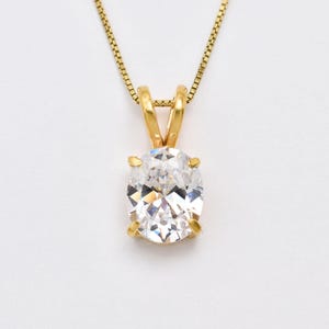 May include: A gold pendant necklace featuring a single oval-cut clear gemstone held in a four-prong setting. The pendant is suspended from a delicate gold box chain, showcasing a classic design with a polished metal finish and faceted stone.