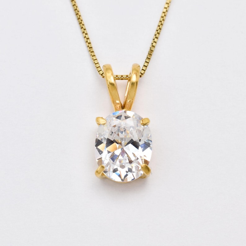 May include: A gold pendant necklace featuring a single oval-cut clear gemstone held in a four-prong setting. The pendant is suspended from a delicate gold box chain, showcasing a classic design with a polished metal finish and faceted stone.
