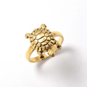 Op de afbeelding: Een goudkleurige ring met een gedetailleerd schildpadontwerp. De schild van de schildpad en de ledematen zijn ingewikkeld gebeeldhouwd, waarbij de ringband soepel aansluit op het lichaam van de schildpad. De ring staat tegen een witte achtergrond.