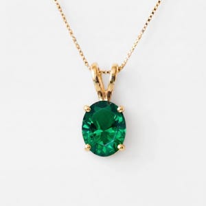 May include: A gold necklace featuring an oval-cut, deep green gemstone pendant held by a four-prong setting. The pendant is suspended from a delicate gold chain, showcasing a faceted surface that reflects light across the stone's vibrant green hue.