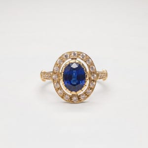 Op de afbeelding: Een gouden ring met een centrale ovaal geslepen blauwe saffier, omringd door een halo van kleine, heldere, rond geslepen diamanten. De zetting is gemaakt van geelgoud, met extra kleine diamanten ingelegd langs de zijkanten van de ringband.