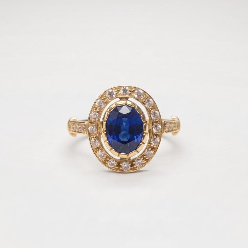 Op de afbeelding: Een gouden ring met een centrale ovaal geslepen blauwe saffier, omringd door een halo van kleine, heldere, rond geslepen diamanten. De zetting is gemaakt van geelgoud, met extra kleine diamanten ingelegd langs de zijkanten van de ringband.