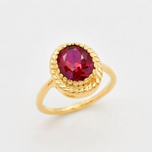 May include: A gold ring featuring a central oval-cut deep red gemstone. The stone is set within a textured, rope-style gold bezel frame. The band is a smooth, polished gold metal, creating a cohesive look for this piece of jewellery.