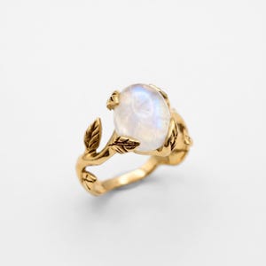 May include: A gold ring with an oval, opalescent gemstone. The ring's design incorporates a nature theme, with leaves and branches forming the band. The gemstone is secured by four prongs. The ring has a polished, shiny finish. The gemstone is approximately 1.5 cm in length.