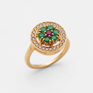 Puede incluir: Un anillo de oro con un diseño floral. El centro del anillo tiene una piedra preciosa roja rodeada de piedras preciosas verdes. El anillo exterior está incrustado con pequeñas piedras preciosas claras.