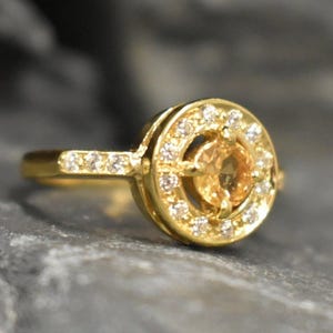 May include: A gold ring with a round, clear gemstone surrounded by smaller clear gemstones. The ring has a simple band.