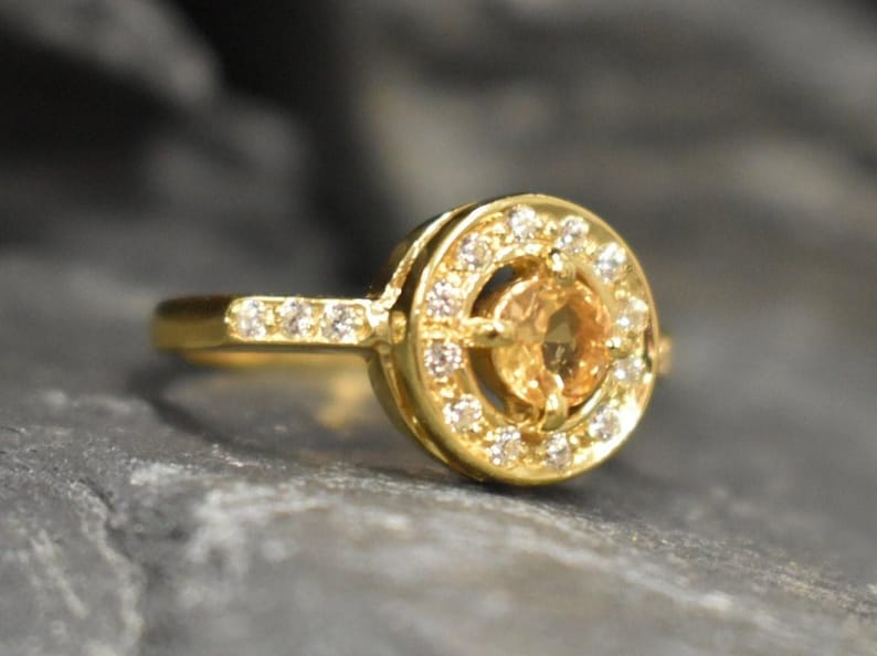 May include: A gold ring with a round, clear gemstone surrounded by smaller clear gemstones. The ring has a simple band.