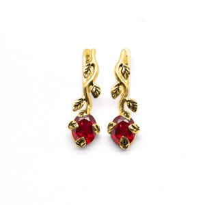 Peut inclure: Une paire de boucles d'oreilles dorées avec un motif de feuilles. Chaque boucle d'oreille présente une pierre précieuse rouge de forme ovale en bas. Les boucles d'oreilles ont une esthétique inspirée de la nature.