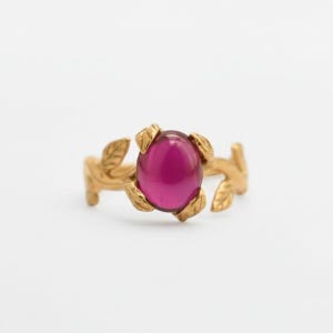 May include: A gold ring with a large, oval, magenta gemstone. The ring's band has a leaf design, giving it an organic feel. The magenta gemstone is the central feature, set in a classic, elegant style. A piece of jewellery.