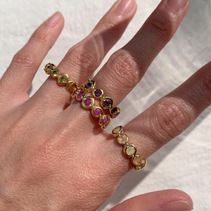 Puede incluir: Tres anillos de oro con piedras preciosas de colores. El anillo superior tiene piedras verdes, amarillas y naranjas. El anillo del medio tiene piedras rosas y moradas. El anillo inferior tiene piedras blancas.