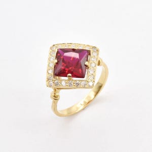 May include: A gold ring with a square-shaped red gemstone surrounded by a halo of smaller white gemstones.