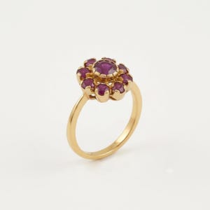 Può includere: Un anello in oro con una pietra preziosa centrale di colore rosa intenso a taglio rotondo, circondata da un gruppo di otto pietre rosa più piccole abbinate a taglio rotondo disposte a fiore. Il gambo è in metallo dorato liscio e lucido.