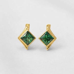 Puede incluir: Un par de pendientes dorados con piedras preciosas verde esmeralda de corte cuadrado. Las piedras están engastadas en un marco dorado con un diseño curvo único. Los pendientes se muestran sobre un fondo blanco.