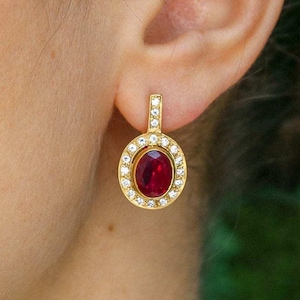 Op de afbeelding: Gouden oorbellen met een rode ovale edelsteen omringd door een halo van kleinere heldere edelstenen.