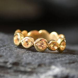 Op de afbeelding: Een gouden ring met een reeks ronde, perzik-gekleurde edelstenen in een herhalend patroon. De ring wordt gepresenteerd op een donkere, getextureerde ondergrond, die de warme tinten en het ingewikkelde ontwerp benadrukt. De band van de ring heeft een geschulpte rand.