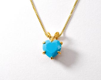 May include: A gold-colored necklace with a heart-shaped, light blue pendant. The pendant is faceted and held in place by gold prongs. The chain is a delicate, gold-colored box chain. The necklace is displayed on a white background.