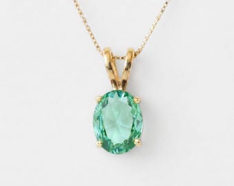May include: A gold necklace featuring an oval-cut light green gemstone pendant held by a four-prong setting. The pendant is suspended from a delicate gold chain that connects to the top of the setting with a simple gold bail.