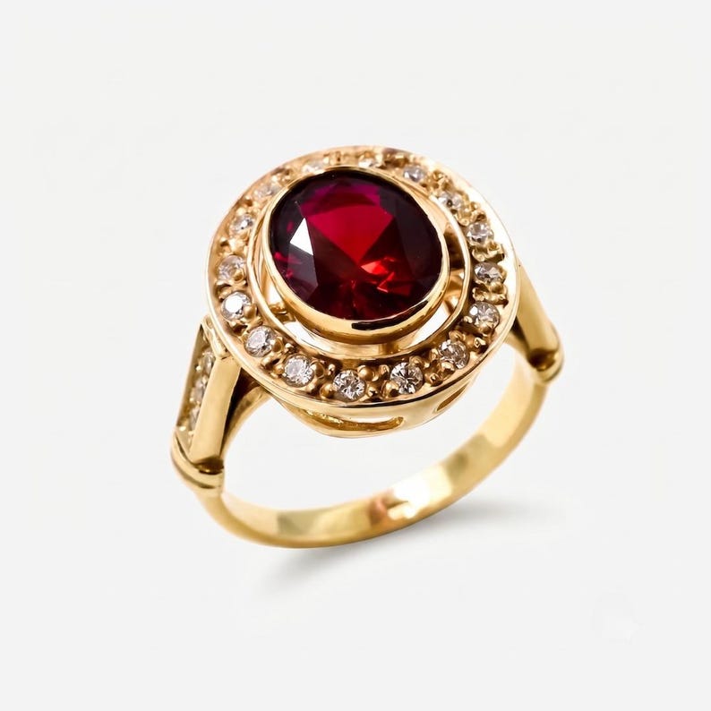May include: A gold ring featuring a large, oval-cut deep red gemstone at the center, surrounded by a halo of small, clear round-cut stones. The band is crafted from polished yellow gold and includes additional small stones along the shoulders.