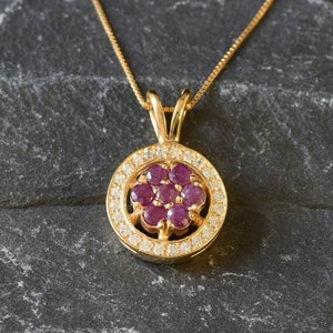 May include: A gold pendant necklace featuring a round ruby and diamond cluster. The pendant is set in a gold circle with a halo of diamonds.