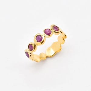 Puede incluir: Un anillo de banda dorada con cinco piedras preciosas redondas de color magenta, engastadas en bisel a lo largo de la mitad superior. El metal tiene un acabado pulido y la banda presenta un diseño de borde festoneado y orgánico.