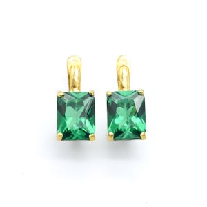 May include: A pair of gold earrings with emerald green square-shaped gemstones. The earrings have a simple, elegant design.