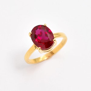 Puede incluir: Un anillo de oro con una gran piedra preciosa de color rojo intenso y corte ovalado, montada en un engaste de cuatro garras. La banda es lisa y pulida, mostrando un tono de oro amarillo brillante que complementa la piedra roja facetada.