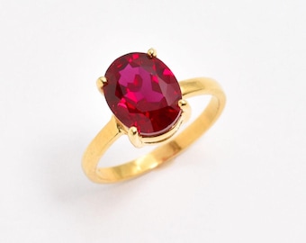 Op de afbeelding: Een gouden ring met een grote, ovaal geslepen, dieprode edelsteen in een zetting met vier pootjes. De ringband is glad en gepolijst en heeft een heldere geelgouden kleur die de levendige, gefacetteerde rode steen in het midden accentueert.