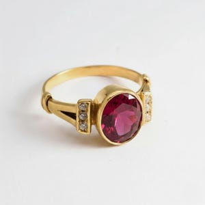 May include: A gold ring featuring a large, oval-cut, deep pink gemstone set in the centre. The band includes two vertical accents on each side, each embedded with three small, clear, round-cut stones that sparkle against the polished gold metal.