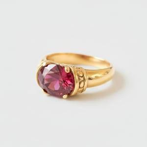 Pode incluir: Um anel de ouro com uma grande pedra preciosa rosa-escura de corte oval numa cravação de garras. A banda é feita de ouro amarelo polido e apresenta um padrão decorativo texturado de pequenos círculos nos lados perto da pedra central.