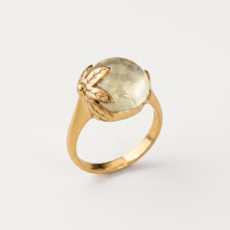 Puede incluir: Un anillo de oro con una piedra preciosa cabuj&oacute;n transl&uacute;cida de color verde p&aacute;lido engastada en una estructura de metal. El engaste presenta un motivo esculpido en forma de hoja en un lado, que se extiende desde la banda para sostener la piedra.