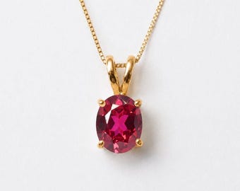 May include: A gold pendant necklace featuring an oval-cut, deep pink gemstone held by a four-prong setting. The pendant is suspended from a delicate gold chain, showcasing a polished metal finish and a vibrant, faceted stone center piece.