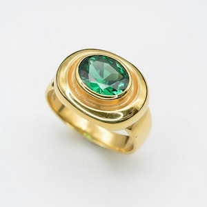 May include: A gold ring with a large, oval green gemstone. The gemstone is set in a gold bezel, and the ring has a smooth, polished band. The ring is displayed against a white backdrop.