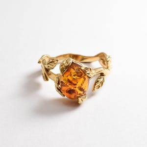 May include: A gold-colored ring featuring a large, oval amber stone. The ring's band is designed with a leaf and vine motif, creating a nature-inspired aesthetic. The amber stone displays a warm, golden-orange hue, adding a touch of elegance to the jewelry.