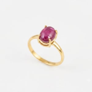 Peut inclure: Une bague en or ornée d'une pierre précieuse rose foncé de forme ovale, taillée en cabochon et fixée par quatre griffes. L'anneau est lisse et poli, présentant un design simple qui met en valeur la couleur vive et la surface arrondie de la pierre.