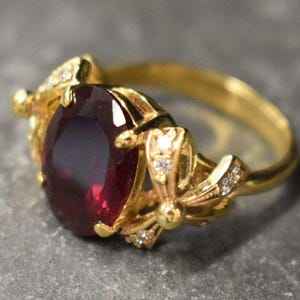 May include: A gold ring with a large, oval-shaped, dark red gemstone surrounded by smaller, clear gemstones. The ring has a delicate, floral design.