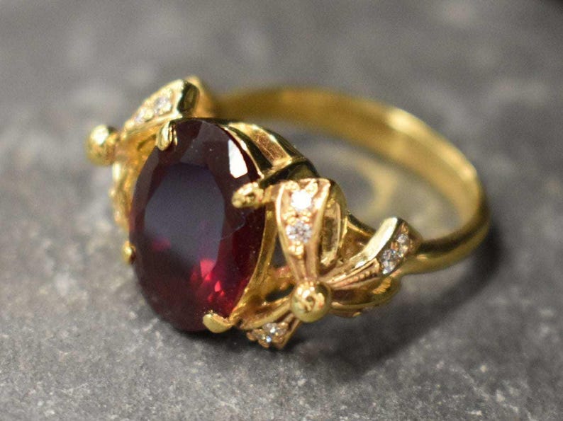 May include: A gold ring with a large, oval-shaped, dark red gemstone surrounded by smaller, clear gemstones. The ring has a delicate, floral design.