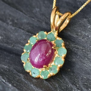 Può includere: Una collana con un ciondolo in oro con una grande pietra di rubino ovale circondata da piccole pietre di smeraldo verde.