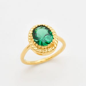 May include: A gold ring with an oval, emerald-green gemstone. The gemstone is set in a textured, rope-like border. The ring's band is a simple, polished gold. The ring is set against a white background.