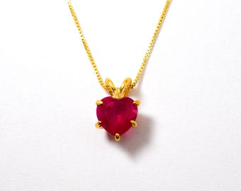 May include: A gold-colored necklace with a heart-shaped, ruby-red pendant. The pendant is held by a gold setting and hangs from a delicate gold chain. The necklace is displayed against a white background.