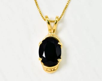 Puede incluir: Un collar dorado con un colgante con una gran piedra preciosa negra de forma ovalada. El colgante está engastado en un marco de metal dorado y cuelga de una delicada cadena dorada. La joya se exhibe sobre un fondo blanco.