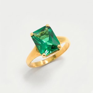 May include: A gold ring featuring a large, emerald-cut green gemstone. The ring has a simple, elegant design with a polished gold band and four prongs securing the gemstone. The gemstone is a vibrant green color.