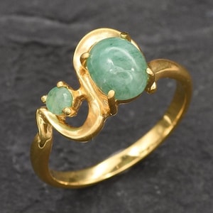 Puede incluir: Un anillo de oro con dos piedras preciosas verdes de forma ovalada. El anillo tiene un dise&ntilde;o &uacute;nico y curvo.