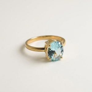 Puede incluir: Un anillo de oro con una piedra preciosa de color azul claro en talla ovalada, sujeta por un engaste de cuatro garras. La banda es lisa y pulida, mostrando un diseño clásico que resalta la claridad y el tono azul pálido de la piedra.