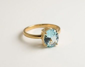 Puede incluir: Un anillo de oro con una piedra preciosa de color azul claro en talla ovalada, sujeta por un engaste de cuatro garras. La banda es lisa y pulida, mostrando un diseño clásico que resalta la claridad y el tono azul pálido de la piedra.