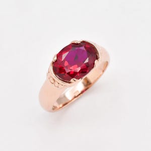 Peut inclure: Une bague en or rose ornée d'une grande pierre précieuse rouge foncé de taille ovale, fixée par des griffes. L'anneau présente une finition lisse et polie avec un motif circulaire décoratif gravé sur les côtés près de la pierre centrale.