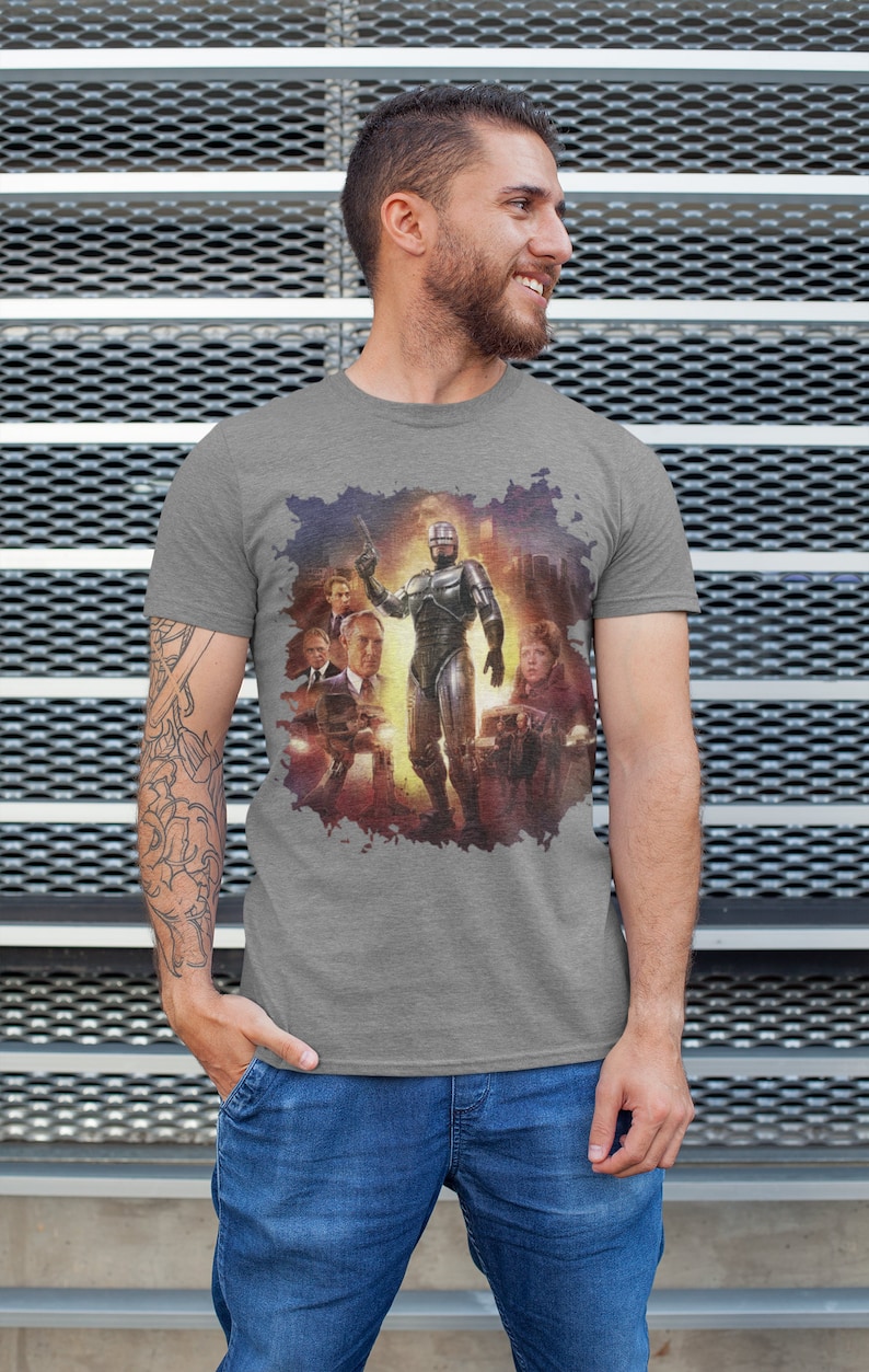 May include: A heather grey t-shirt with a graphic print of a scene from the movie Robocop. The graphic features Robocop standing in the middle of the scene with other characters from the movie surrounding him.