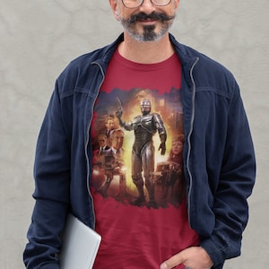 May include: A man wearing a red t-shirt with a graphic of Robocop and other characters from the movie. He is also wearing a blue jacket and jeans. He is holding a laptop computer in his left hand.
